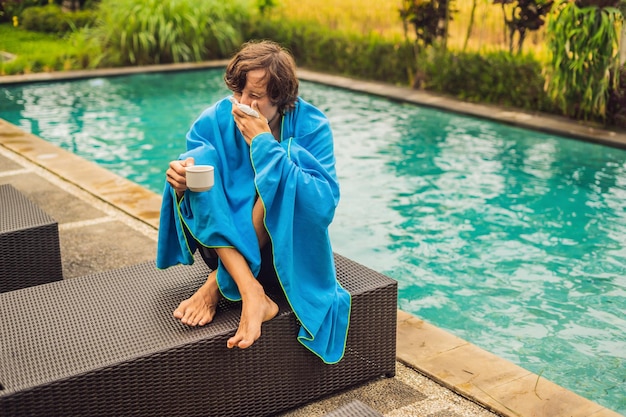 Zieke man reiziger De man is verkouden op vakantie zit verdrietig aan het zwembad thee te drinken en snuit zijn neus in een servet Zijn zoon is gezond en zwemt in het zwembad Reisverzekeringsconcept