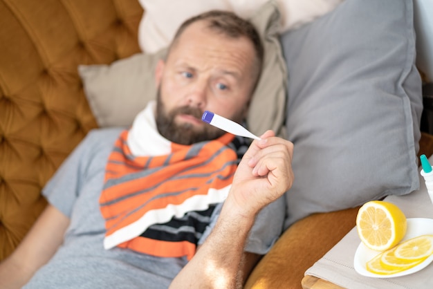 Zieke mens die op bank ligt die zijn temperatuur thuis in de woonkamer controleert