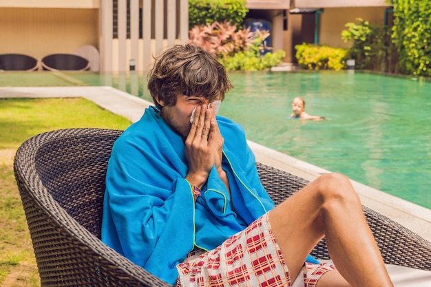 Zieke mensenreiziger. De man is verkouden op vakantie, zit bedroefd aan het zwembad thee te drinken en snuit zijn neus in een servet.