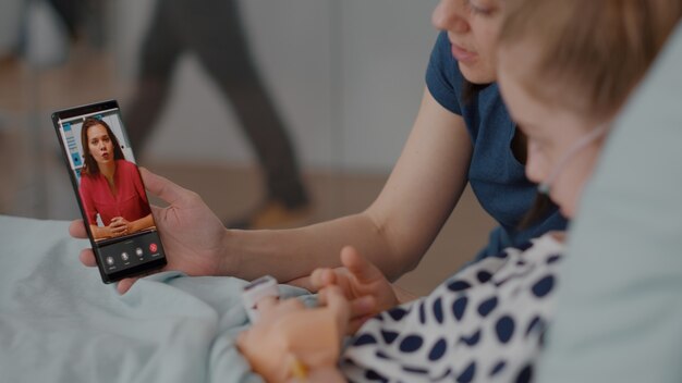 Zieke patiënt rust in bed met moeder die vriend op afstand begroet tijdens online videogesprek