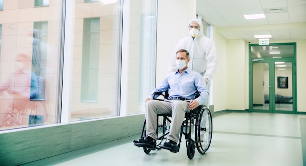 Zieke senior man met beschermend veiligheidsmasker op gezicht in een rolstoel en een zelfverzekerde arts in het medische masker tijdens het vervoer in het ziekenhuis.