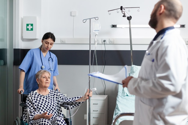 Zieke senior vrouw zittend in een rolstoel met infuuszak bevestigd op arm