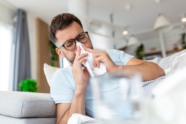 Zieke trieste man met bril zit thuis op de bank lijdt aan loopneus griepziekte coronavirus pandemie covid epidemie niest Onwel man voelt zich slecht koorts virus ziektesymptomen binnen