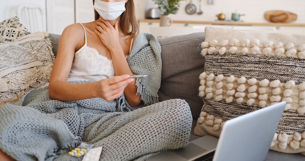 Zieke vrouw die het masker van de gezichtsbescherming draagt, op laag met laptop zit en thermometer en pillen houdt tijdens huis quarantaine zelfisolatie.