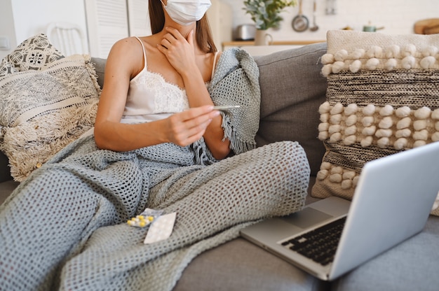 Zieke vrouw die het masker van de gezichtsbescherming draagt, op laag met laptop zit en thermometer en pillen houdt tijdens huis quarantaine zelfisolatie.
