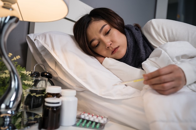 Zieke vrouw die thermometer gebruikt om haar temperatuur en haar hoofdpijn in bed te controleren