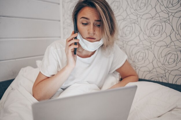 Zieke vrouw in beschermend gezichtsmasker dat in bed ligt, smartphone spreekt en aan laptop werkt tijdens de isolatie van het huisquarantaine Covid-19 pandemisch Corona-virus. Afstand online werk vanuit huis concept.