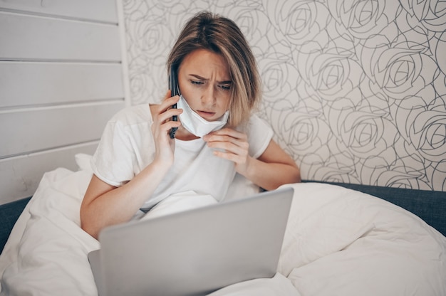 Zieke vrouw in gezichtsmasker die smartphone spreken en aan laptop in bed werken