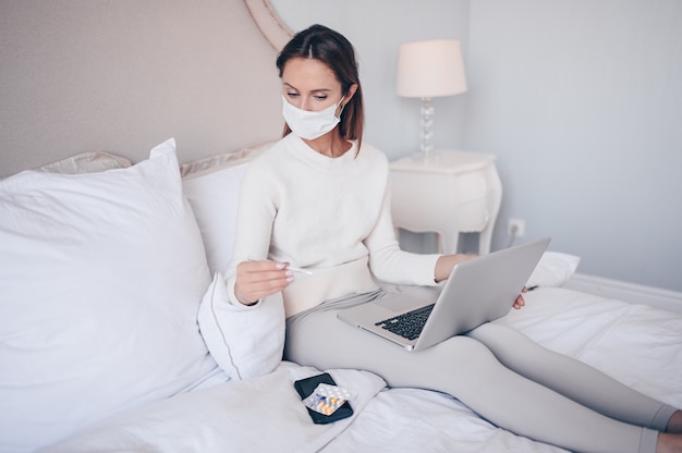 Zieke vrouw in het masker van de gezichtsbescherming die in bed met laptop holdingsthermometer en pillen thuis liggen