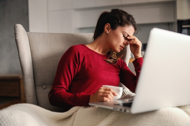 Zieke vrouw zittend in stoel thuis tijdens lockdown met laptop op haar schoot en hoofdpijn.