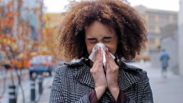 Zieke zieke Afro-Amerikaanse vrouw met krullend haar blaast loopende neus in zakdoek buiten koud