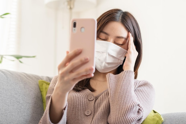 Zieke, zieke aziatische jonge vrouw, meisje met masker met keelpijn, hoofdpijn heeft koorts, griep en gebruikt smartphone-consultatie met arts online, telehealth thuis. Gezondheidszorg persoon op virus seizoensgebonden.