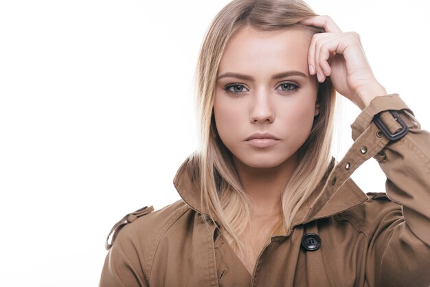 Ziet er mooi uit. Aantrekkelijke jonge vrouw in jas die haar haar aanpast en naar de camera kijkt terwijl ze tegen een witte achtergrond staat