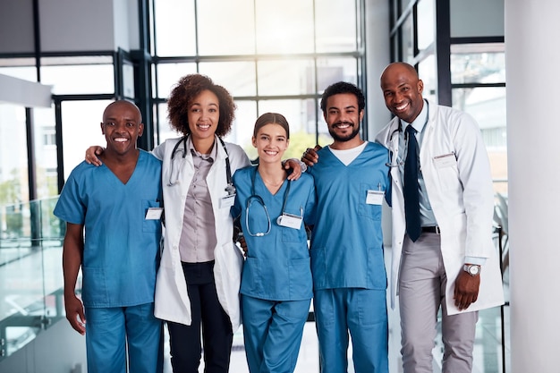 Zij zullen voor uw medische behoeften zorgen Portret van een vrolijke groep artsen die overdag met hun armen om elkaar heen staan in een ziekenhuis