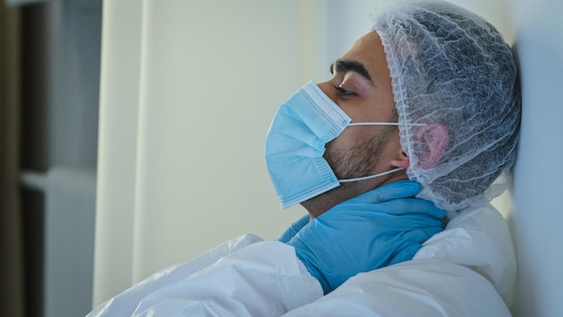 Zijaanzicht arts met masker zittend op de vloer in de kliniek in de buurt van de muur met de handen moe