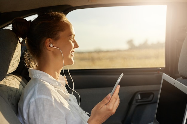 Zijaanzicht close-up portret van zakenvrouw zittend op de achterbank van de auto en werken met laptop luisteren muziek in oortelefoons met mobiele telefoon in de hand kijkend naar raam genietend van zonsondergang