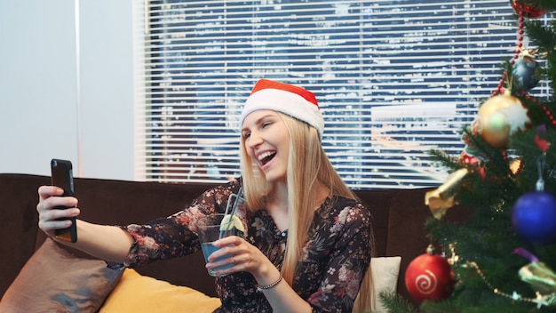 Zijaanzicht die van knappe vrouw selfie in Kerstmanhoed en met drank in haar handen maken