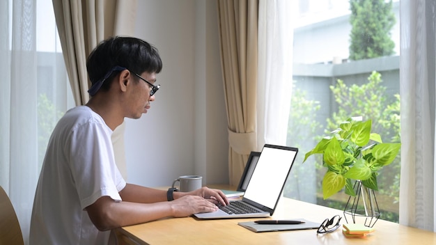 Zijaanzicht jonge aziatische man zit in een comfortabele woonkamer en surft op internet op een laptopcomputer