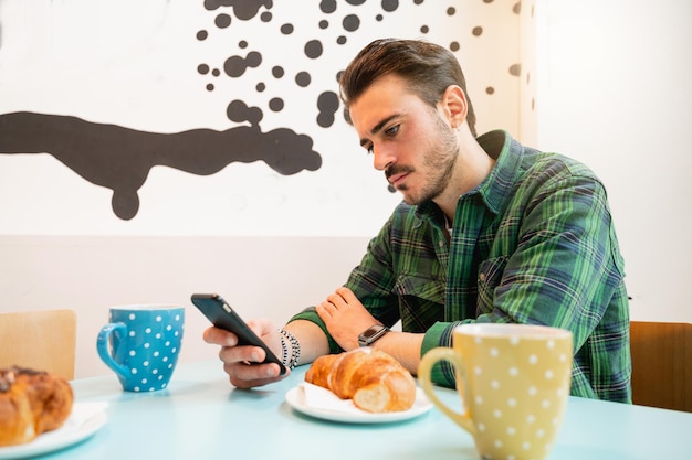 Zijaanzicht jonge hipster man zit aan tafel café met smartphone Mokken en croissants op tafel