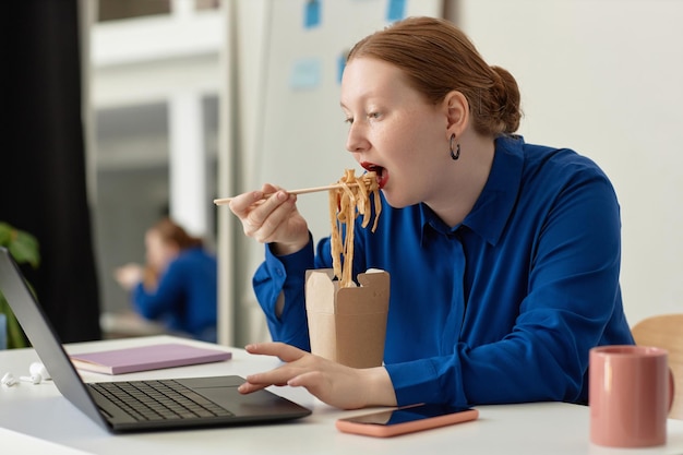 Zijaanzicht jonge vrouw die meeneemnoedels eet op de werkplek op kantoor