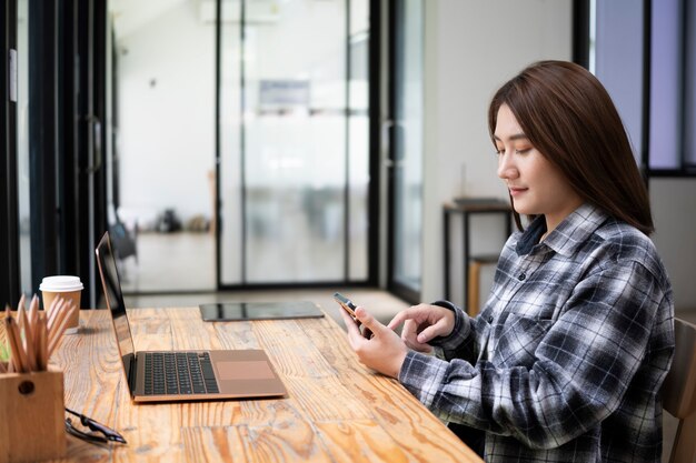 Zijaanzicht jonge vrouw die voor laptop op kantoor zit en mobiele telefoon gebruikt.