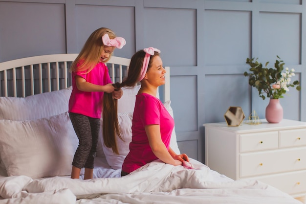 Zijaanzicht klein meisje in pinktshirt staande op het bed en haar moeder kammen Jonge mooie vrouw zittend in bed terwijl haar dochter speelt met haar lange haren schoonheidssalon thuis