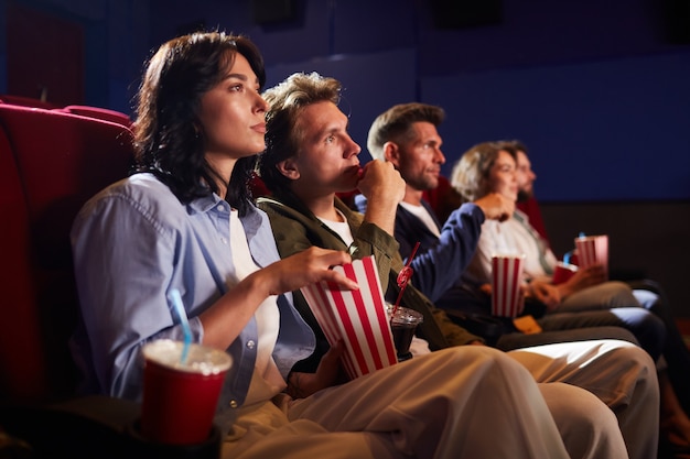 Zijaanzicht op groep vrienden kijken naar film in bioscoop theater en popcorn eten zittend in rij op rode stoelen, kopie ruimte