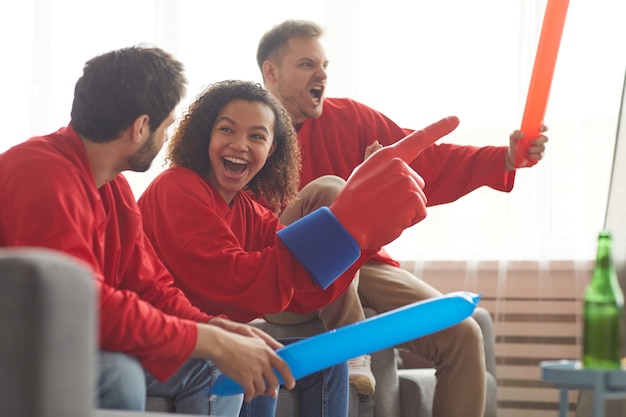 Zijaanzicht op groep vrienden kijken naar sportwedstrijd op Tv thuis en emotioneel juichen terwijl ze rode teamuniformen dragen