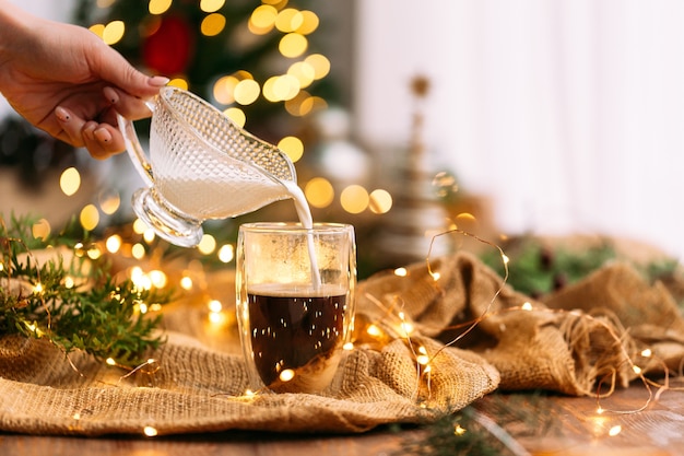 Zijaanzicht op het gieten van melk in een glazen mok koffie op de feestelijke slingerachtergrond