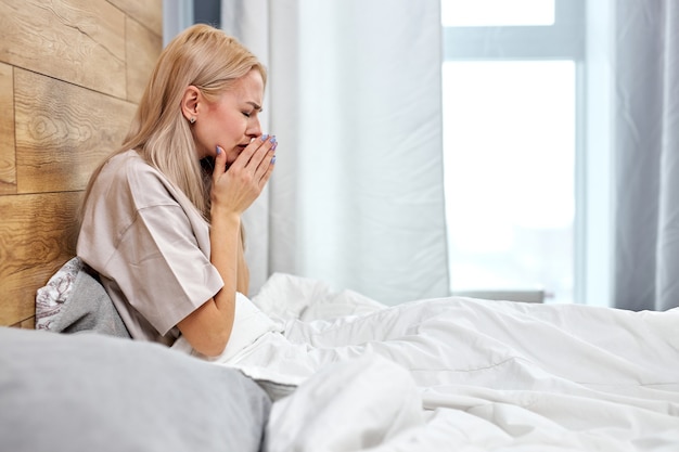 Zijaanzicht op vrouw ziek met griep thuis zittend op bed alleen, met pijn in de keel. zieke blanke vrouw met seizoensgebonden infecties, griepallergie en loopneus. coronavirus (COVID-19