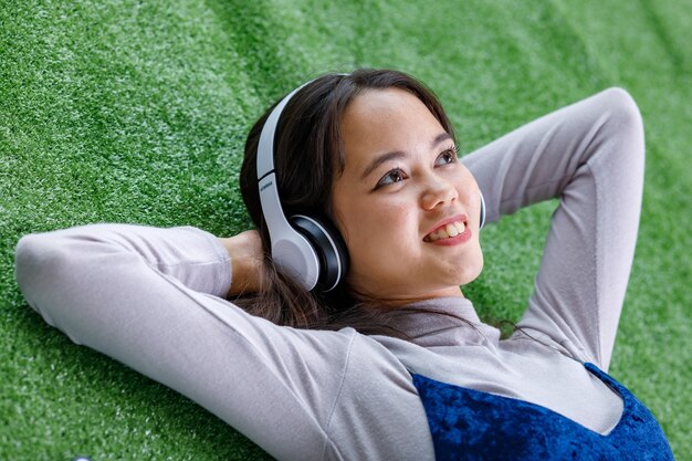 Zijaanzicht portret shot van een schattige lachende jonge Thais-Turkse tiener die geniet van het luisteren naar muziek met een headset. Junior meisje met vrijetijdskleding die op het kunstgras ligt en naar de lucht kijkt
