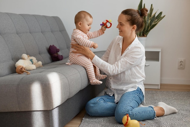 Zijaanzicht portret van blanke vrouw met wit overhemd en spijkerbroek zittend op de bank met haar dochtertje, schattig kind zittend op hoest en opgewonden kijkend.