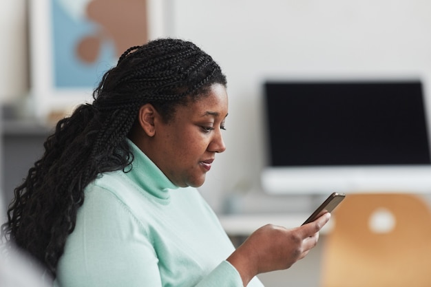 Zijaanzicht portret van bochtige Afro-Amerikaanse vrouw die op het scherm van de smartphone kijkt terwijl ze op internet surft zittend op de bank in een minimaal interieur, kopieer ruimte