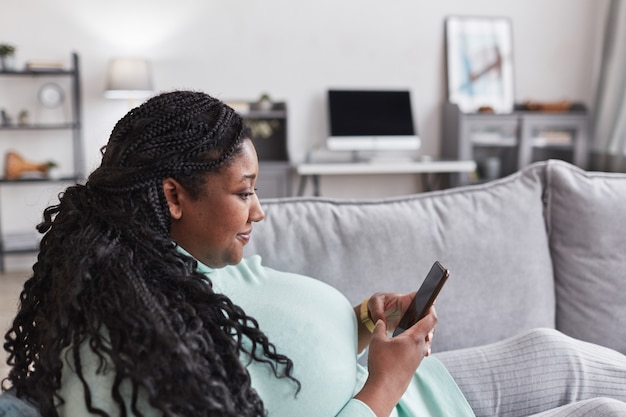 Zijaanzicht portret van bochtige Afro-Amerikaanse vrouw die smartphone gebruikt en glimlacht terwijl ze op de bank ligt en thuis ontspant in een minimaal interieur, kopieer ruimte