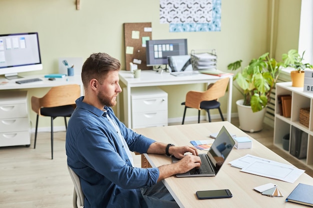 Zijaanzicht portret van een bebaarde jongeman die een laptop gebruikt terwijl hij in een gezellige kantoorruimte werkt