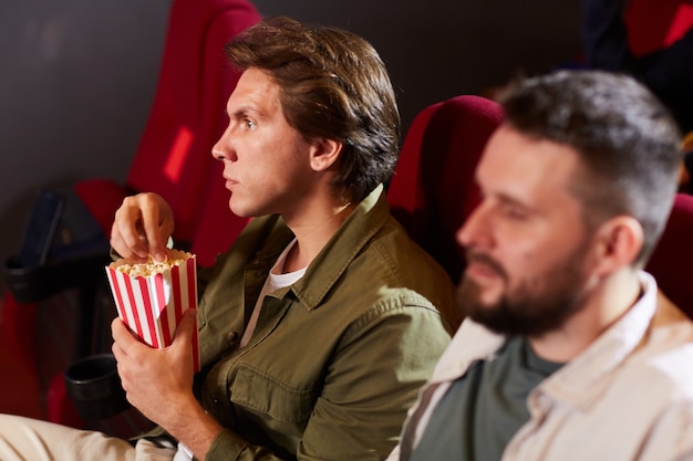 Zijaanzicht portret van een jonge man die popcorn eet tijdens het kijken naar film in de bioscoop met andere mensen op de voorgrond, kopie ruimte