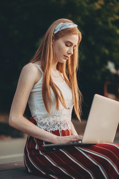 Zijaanzicht portret van een mooie vrouw met rood haar en sproeten kijken naar een laptopscherm zittend op een bankje in het park.