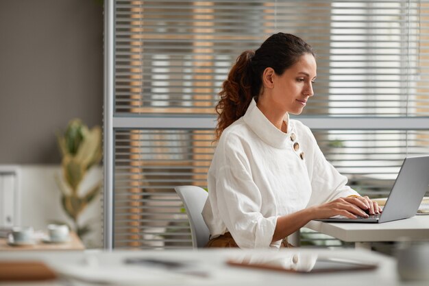 Zijaanzicht portret van elegante succesvolle zakenvrouw met behulp van laptop tijdens het werken aan een bureau in een modern kantoor, kopieer ruimte