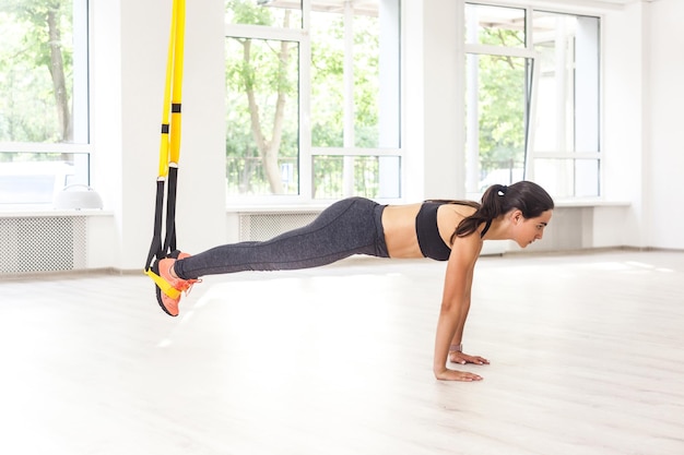 Zijaanzicht portret van jonge gespierde vrouw met zwarte top en legging die op de plank staat, fitnessriemen gebruikt, push-ups doet terwijl de benen aan trx hangen. Binnen, raamachtergrond