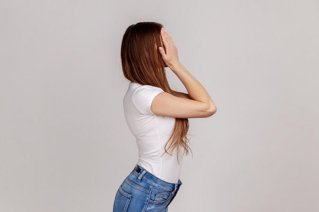 Zijaanzicht portret van jonge volwassen donkerharige vrouw staande die ogen bedekken met de hand weigeren te kijken naar schaamte inhoud dragen witte Tshirt Indoor studio opname geïsoleerd op een grijze achtergrond