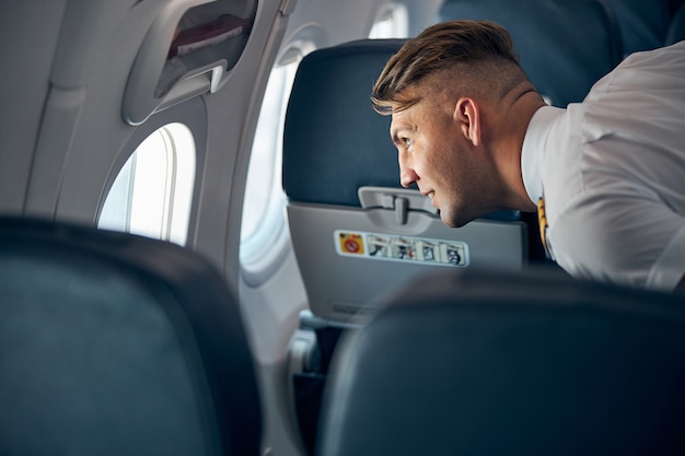 Zijaanzicht portret van knappe man in wit overhemd in de salon van het vliegtuig kijkend naar het raam bij de blauwe passagiersstoelen