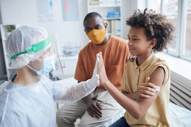 Zijaanzicht portret van lachende Afro-Amerikaanse jongen high five verpleegster na covid-vaccinatie in kliniek of ziekenhuis, kopieer ruimte