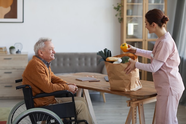 Zijaanzicht portret van lachende senior man in rolstoel kijkend naar vrouwelijke verpleegster die boodschappen, hulp en voedselbezorgingsconcept brengt