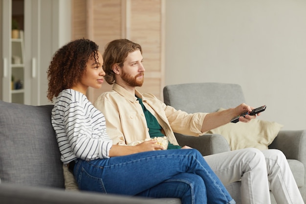 Zijaanzicht portret van moderne mixed-race paar tv kijken thuis terwijl u ontspant op een gezellige bank, focus op kralen man met afstandsbediening