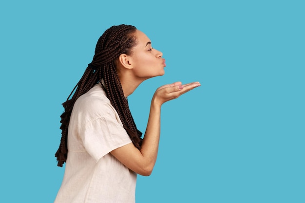 Zijaanzicht portret van romantische vrouw die luchtkus blaast, houdt de lippen gevouwen palm naar mond, stuurt mwah koket, wil haar partner opvrolijken. Indoor studio opname geïsoleerd op blauwe achtergrond.