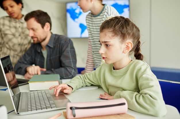 Zijaanzicht portret van schattig schoolmeisje met behulp van laptop in moderne schoolklas met kinderen in backgroun