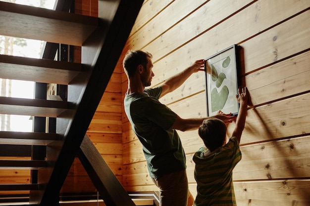 Zijaanzicht portret van vader en zoon die een foto aan de muur hangen in een nieuw huis met achtergrondverlichting