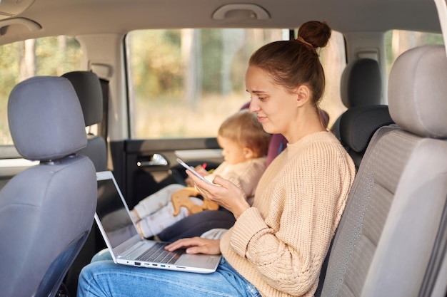 Zijaanzicht portret van verbaasde hulpeloze vrouw die op laptop werkt terwijl ze met haar dochtertje in veiligheidsstoel op de achterbank van de auto zit met behulp van mobiele telefoon met verwarde uitdrukking