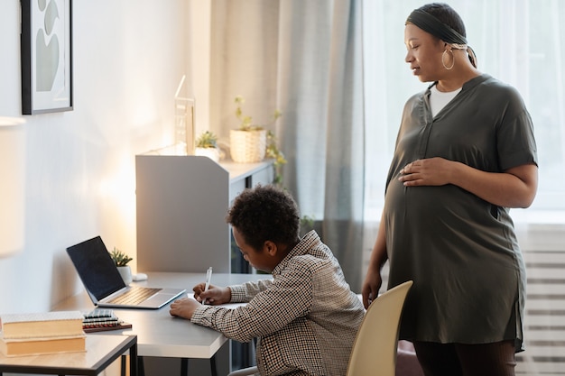 Zijaanzicht portret van zwangere Afro-Amerikaanse vrouw kijken tienerzoon huiswerk, kopieer ruimte