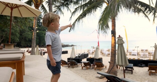 Zijaanzicht van aanbiddelijke kleine jongen in t-shirt en borrels die op zee richten terwijl status dichtbij lanterfanters op strand bij toevlucht